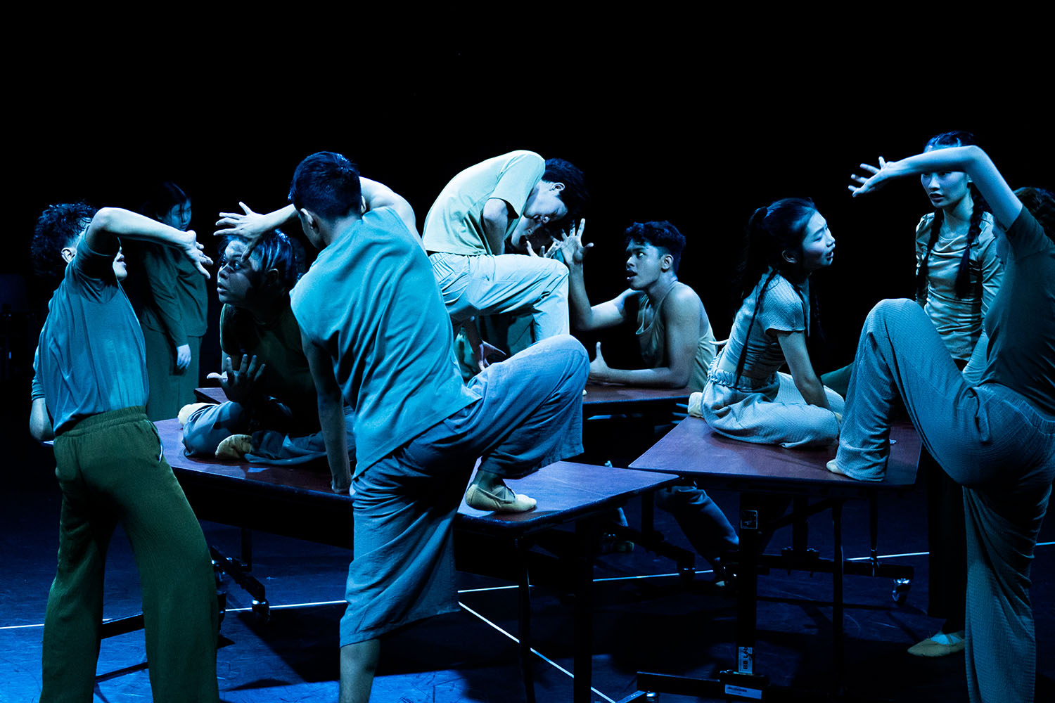 Various performers performing on stage surrounded with long tables under blue light.