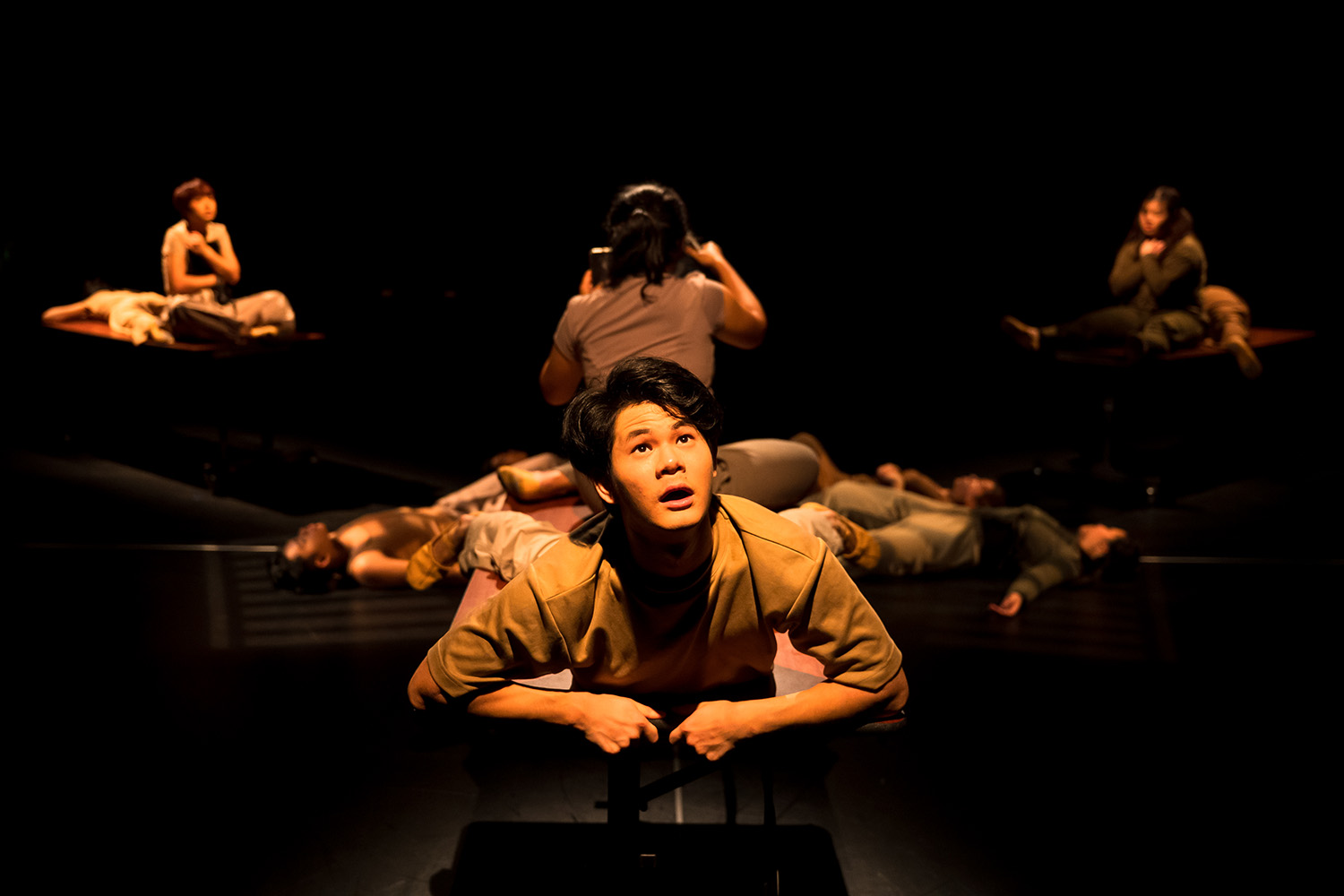 Various performers performing on stage, focus on a male performer centre-stage. Brown-yellow light all over.