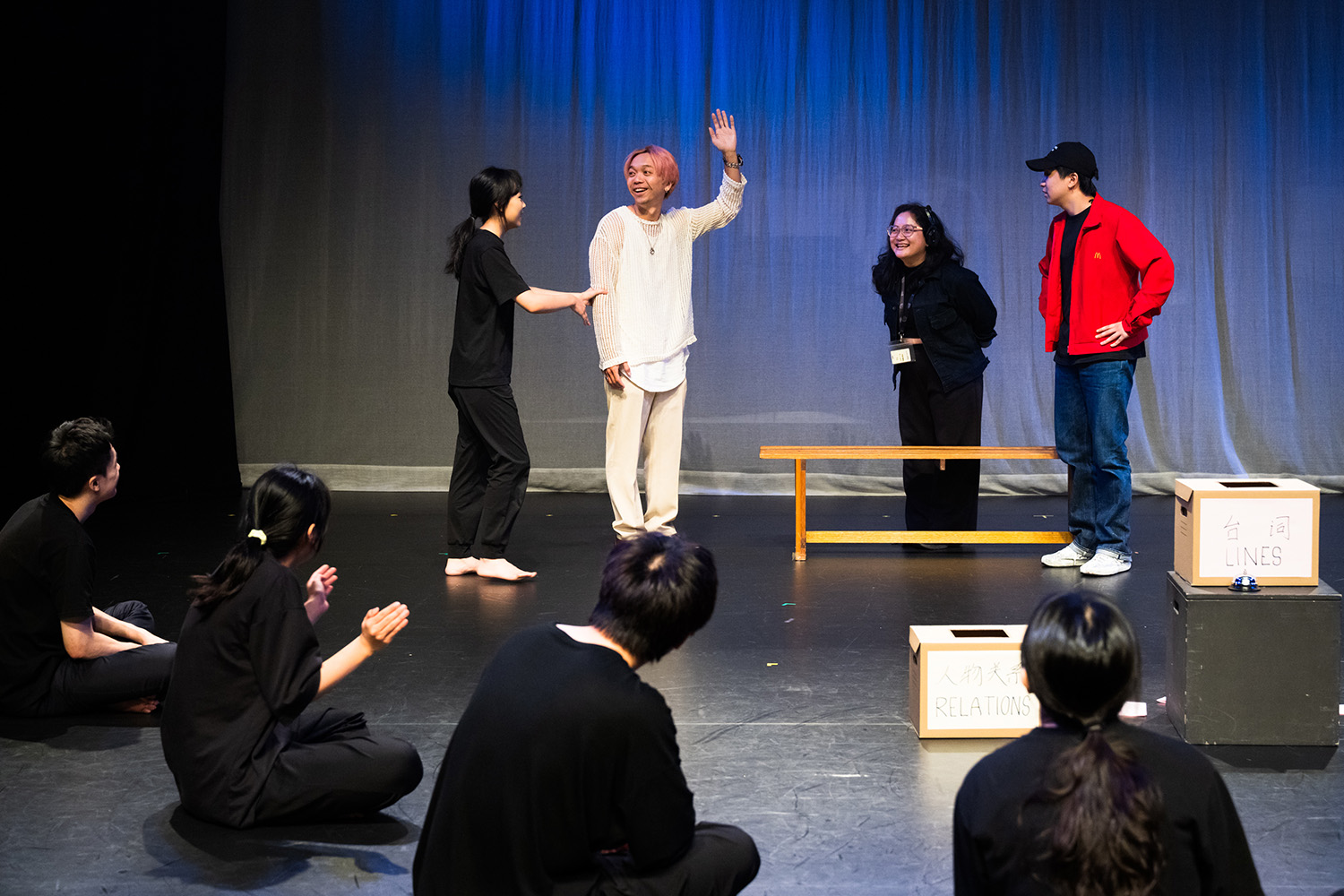 A purple-haired actor in white costume with many other performers wearing black acting on-stage.