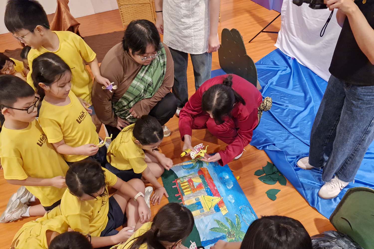 Children, mostly in yellow shirts, joyfully gather around a piece of paper that is a coloured crayon drawing.