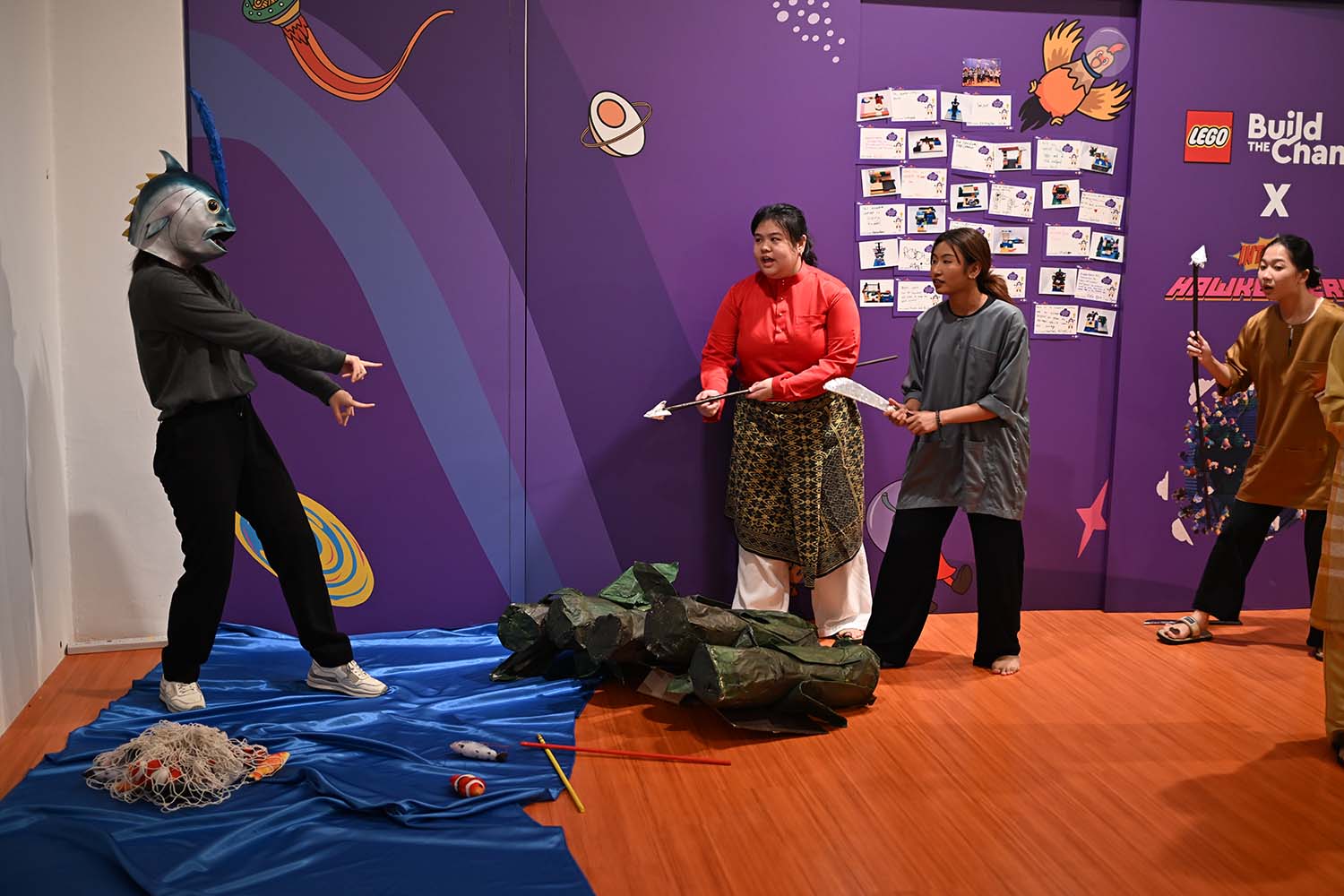 A group of people in traditional Malay costumes stands by a purple wall, interacting with an actor in a fish head mask.