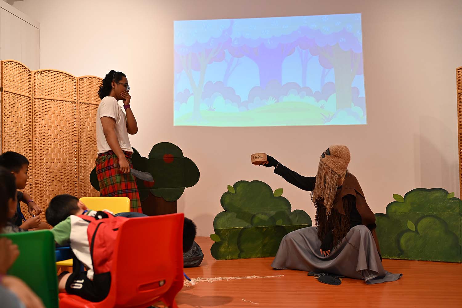 A woman in a headscarf engaging with various kids in a role-playing story-telling session, the background showing a projector with an illustration of a forest.