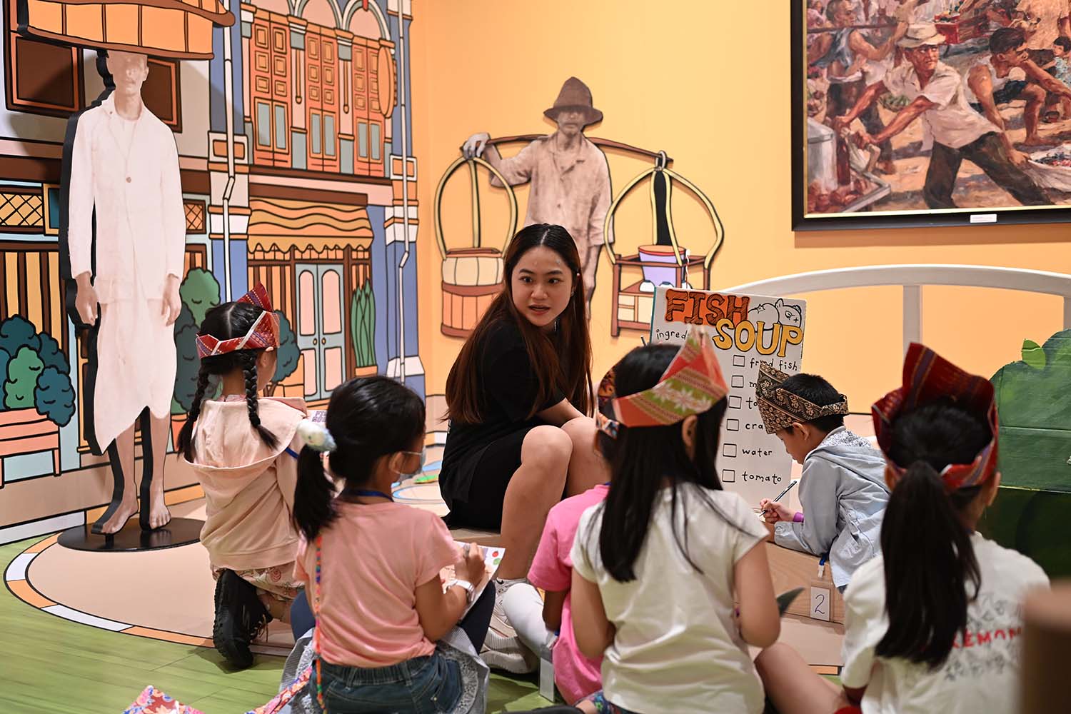A female session conductor carrying ouy a group activity with a group of very young participants, a large write-up sheet with the header reading Fish Soup Recipe.