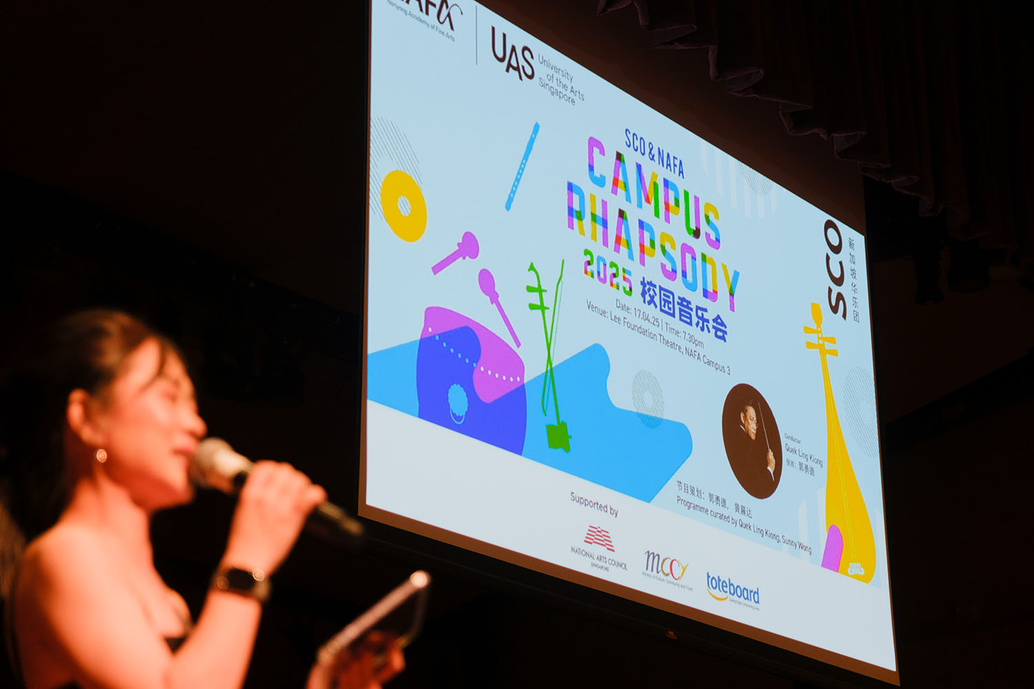 Presenter speaking on-stage with the projector showing Campus Rhapsody 2025 at the Lee Foundation Theatre.