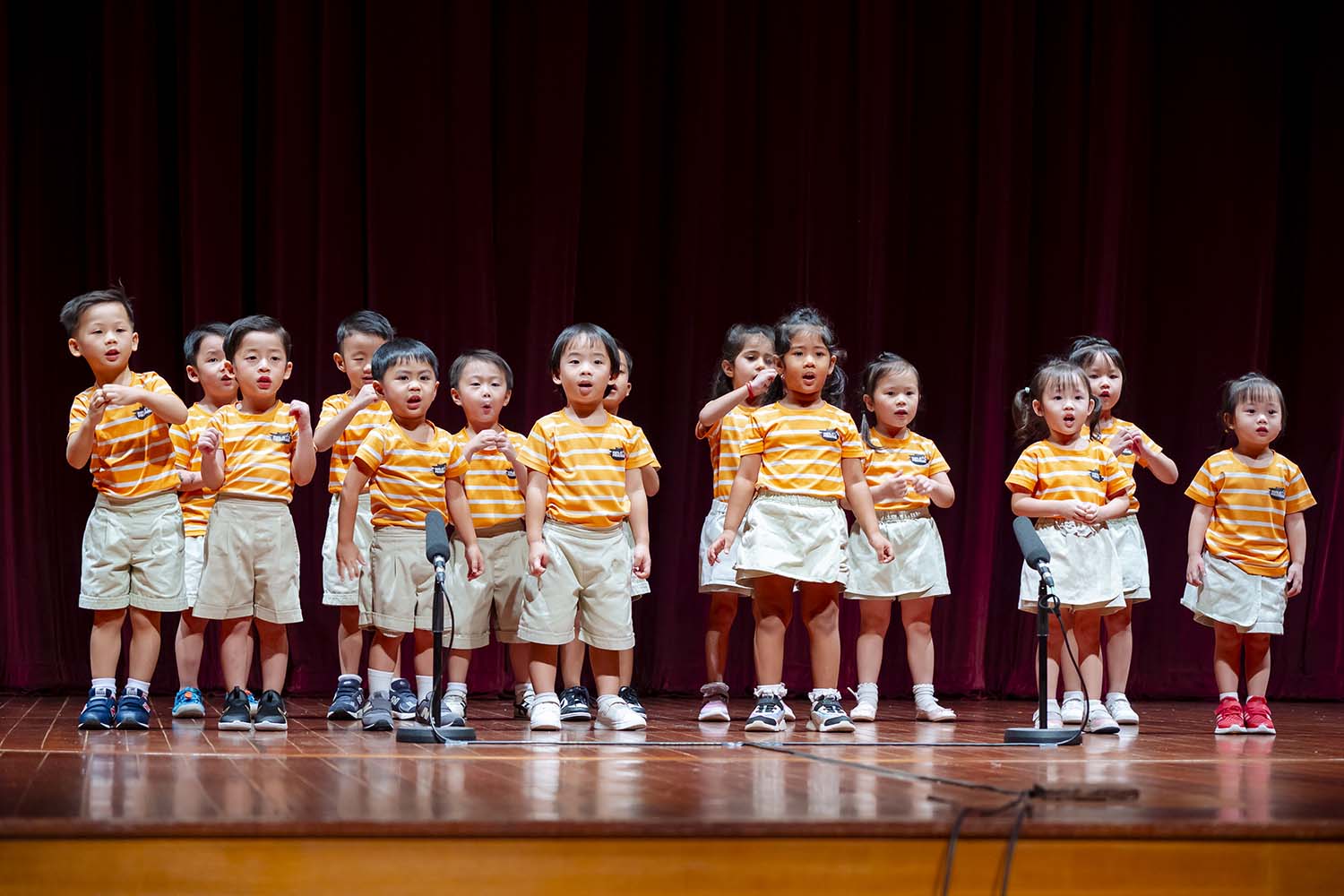 Various preschool K2 young learners in yellow-orange striped t-shirts and shorts on stage.
