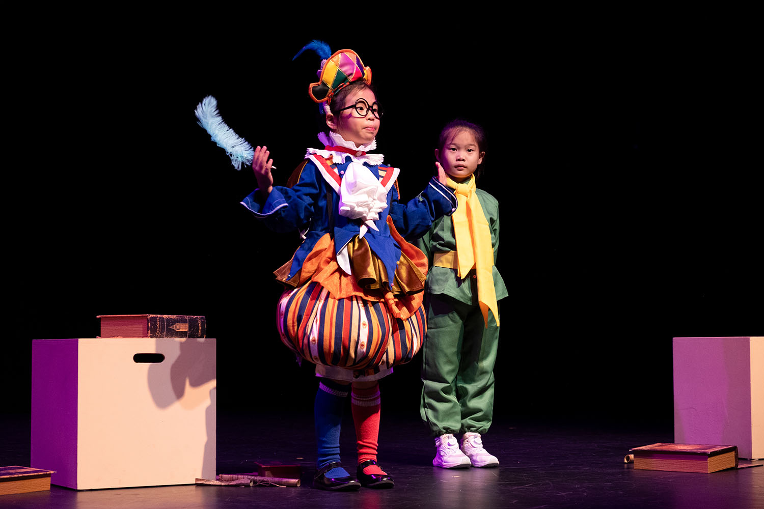 2 Junior Drama performers, the left one wearing an elaborate jester costume, the right one wearing a green traditional costume with a yellow scarf.