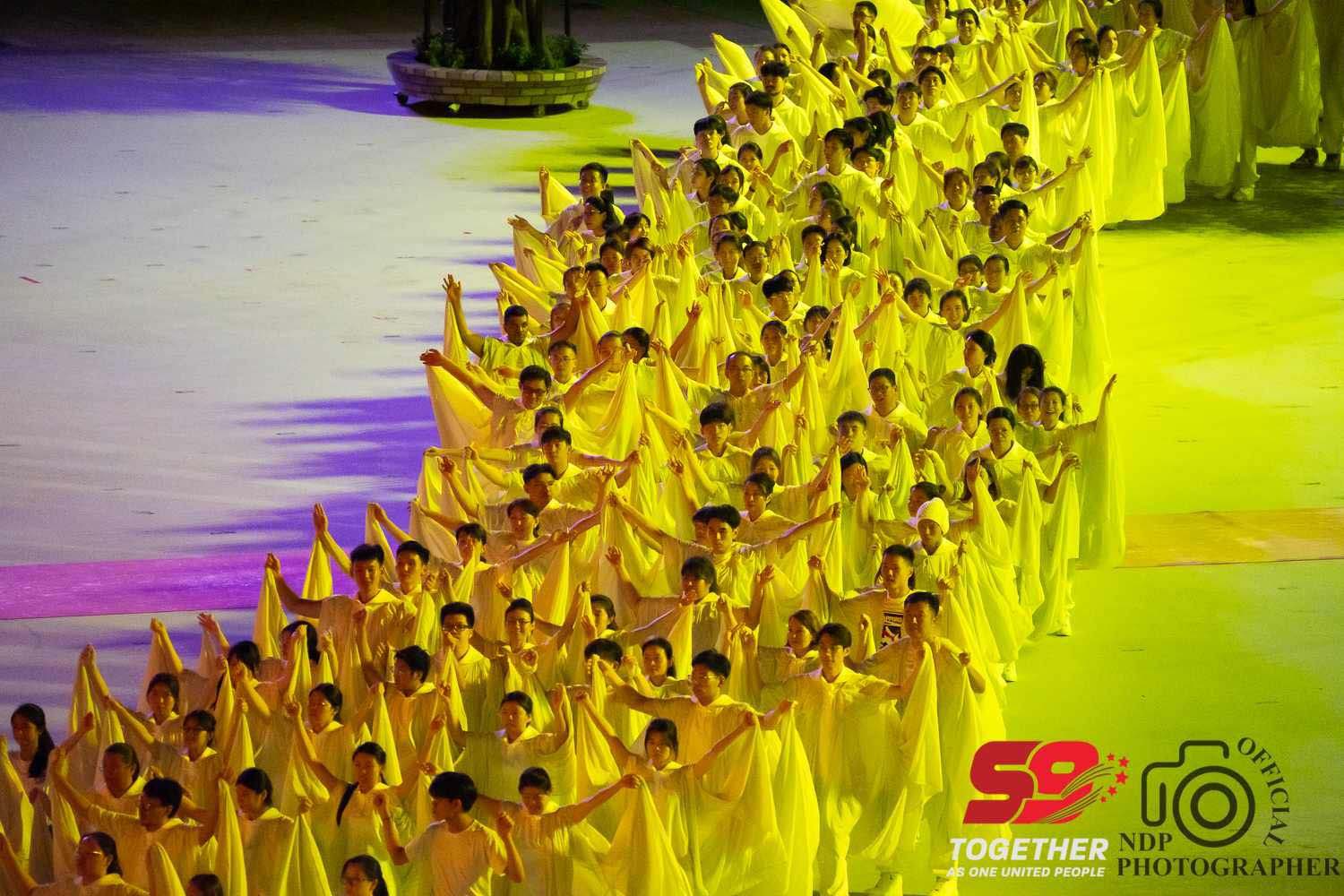 NDP59 various performances in lumiscent yellow suits performing on parade under yellow light at night.