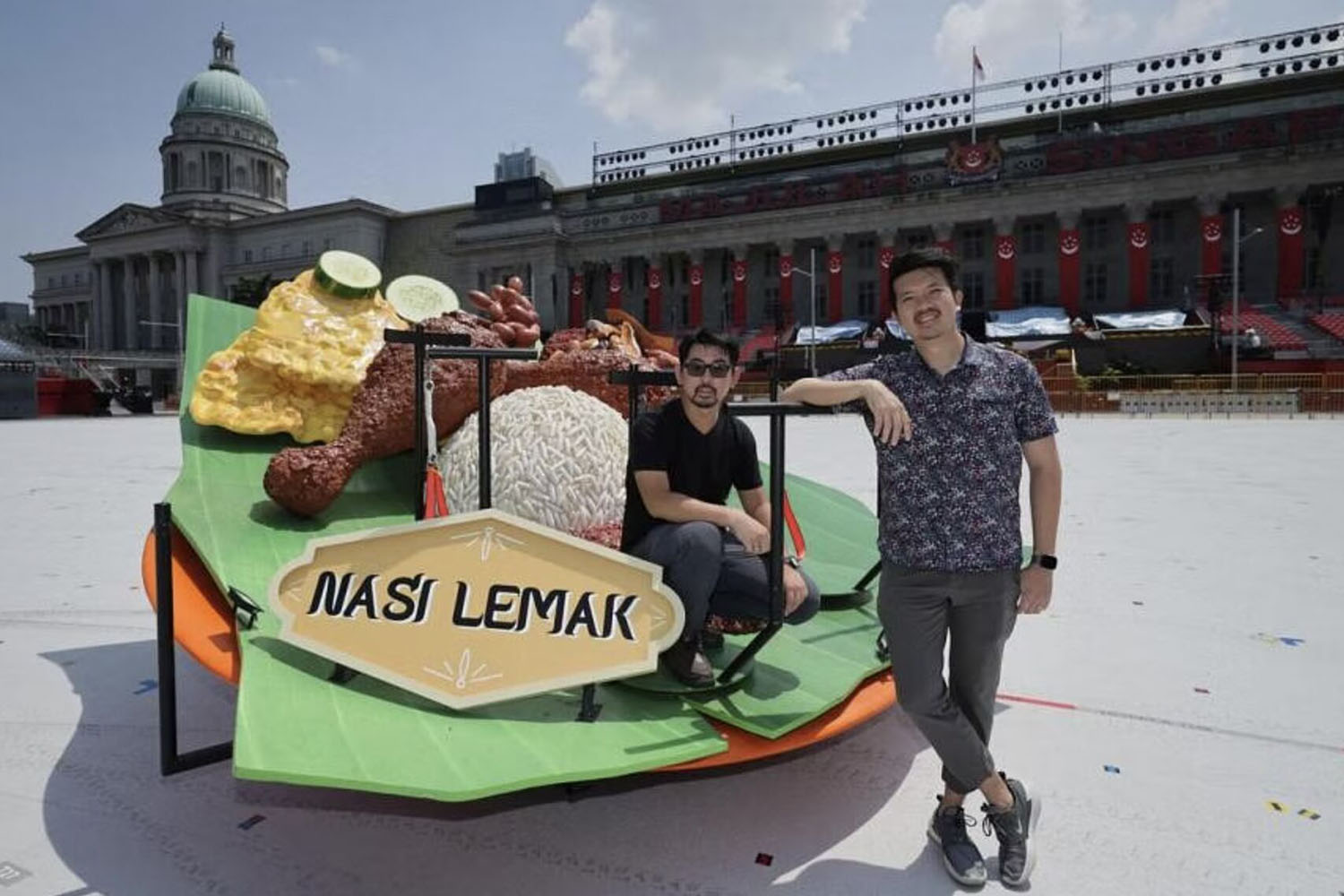 Two persons in casual clothese seated around a mobile Nasi Lemak stage prop on the parade area.